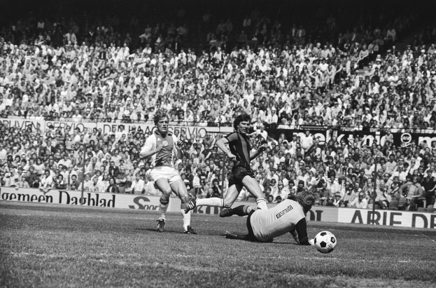 Schwarzes Foto von Männern, die auf einem Feld Fussball spielen, mit Zuschauern im Hintergrund und einer 'Bundesliga 1978-79 Ajax 1-1 Hannover 96' Fahne.