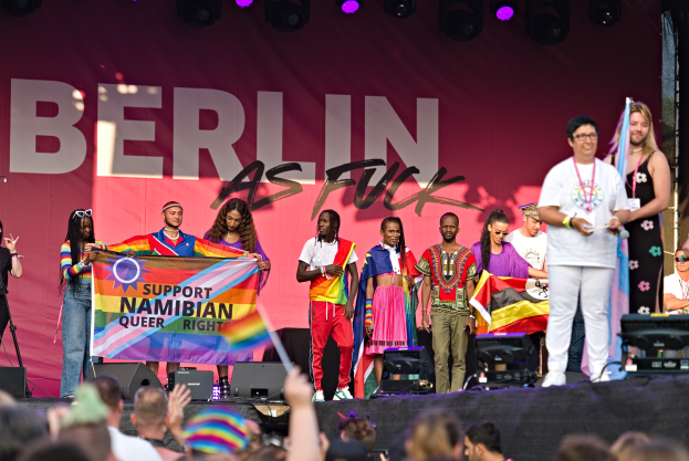Gruppe von Menschen auf einer Bühne mit einem Banner, auf dem 'Berlin as Fuck' steht, mit Lautsprechern, Mikrofonständen und einer Menge unten, die Hände erhebt.
