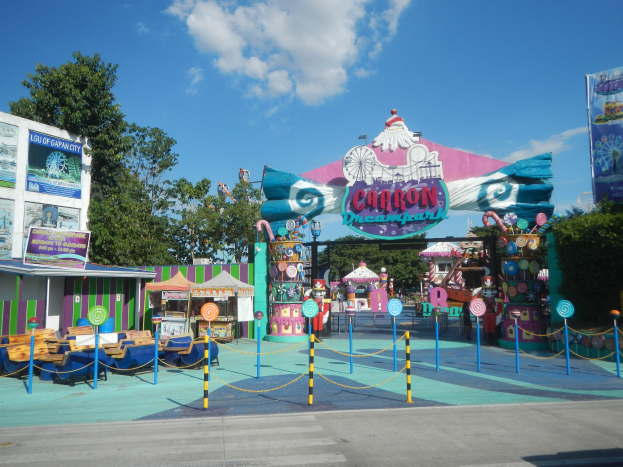 Kirmesfahrgeschäft am Parkeingang mit Straße, Zaun, Buden, Schildern, Bäumen und bewölktem Himmel im Hintergrund.
