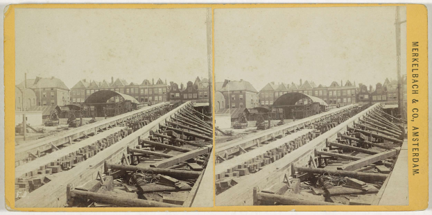 Schwarzes und weißes Foto einer Bahnschiene mit Gebäuden im Hintergrund, Text auf der rechten Seite 'Merkelbach & Co, Co. Amsterdam' und verstreute hölzerne Gegenstände im Vordergrund.