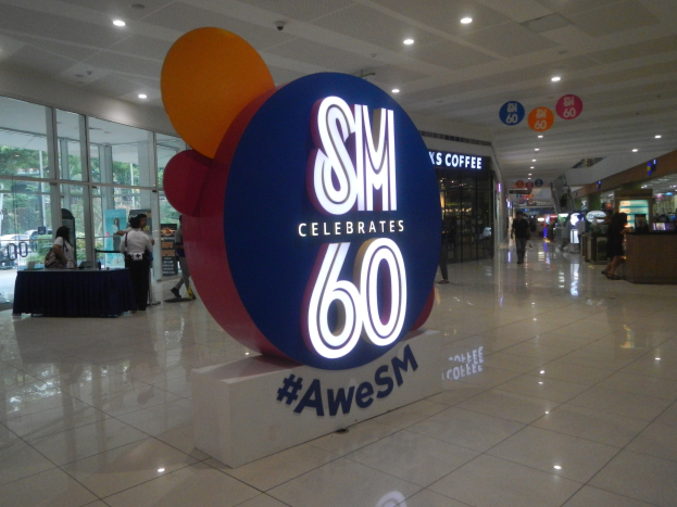 Eine Gruppe von Menschen steht auf einem Einkaufszentrumsboden unter einem "SM feiert 60. Jubiläum"-Schild, mit einem schwarzen-gedeckten Tisch links und Glastüren, Schilden und Deckenleuchten im Hintergrund.