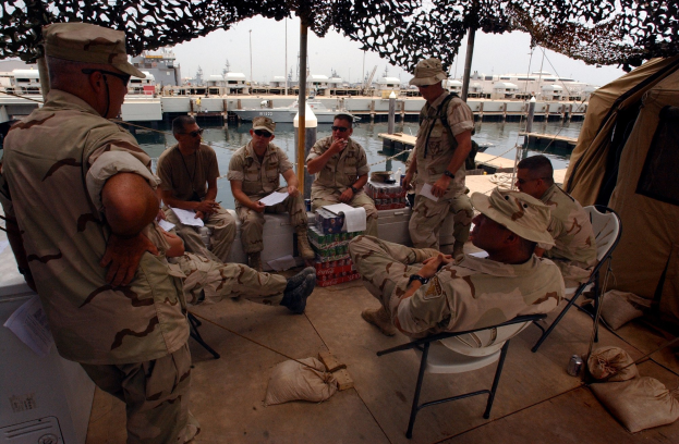 Gruppe von Männern mit Mützen und Schwimmbrillen, die um einen Tisch auf einem Boot sitzen, mit Taschen auf dem Boden und einem Zelt rechts daneben, umgeben von anderen Booten, Pfählen, Gebäuden und einem klaren blauen Himmel.