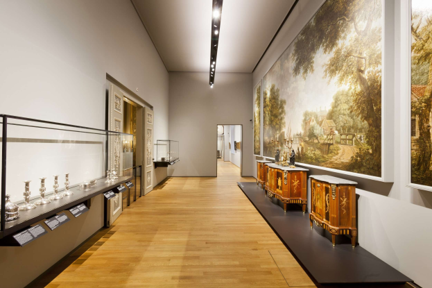 Ein Raum im Museum of Fine Arts, Amsterdam, mit verschiedenen Kunstwerken, darunter Gemälde, Gegenstände auf Tischen, eine Glasvitrine, Informationsschilder, Türen und Deckenleuchten.