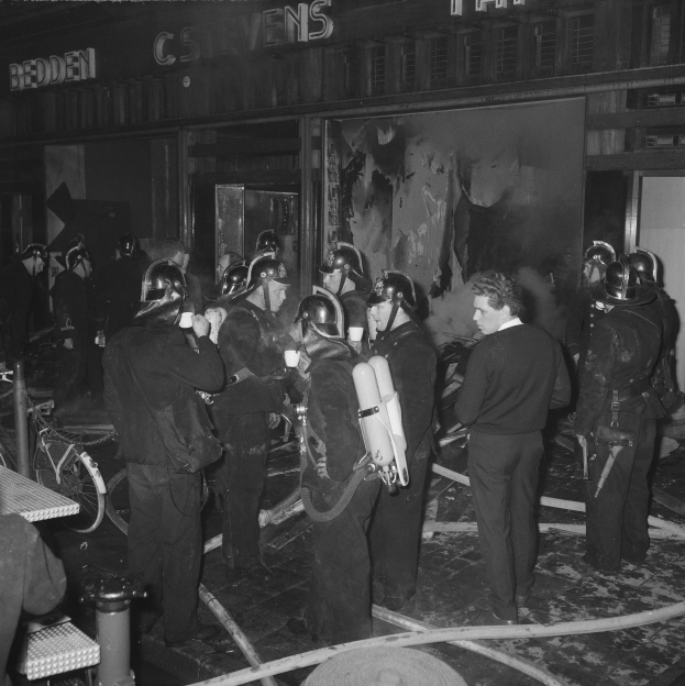 Schwarzes Foto von Feuerwehrleuten in Uniform mit Atemschutz vor einem Gebäude, mit einem Fahrrad und einem Tisch mit Gegenständen in der Nähe.