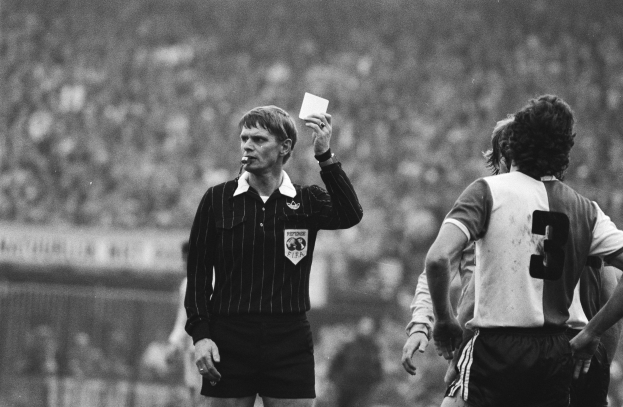 Schwarzes und weißes Bild eines Schiedsrichters, der eine rote Karte in der Luft hält, vor einer Menge von Menschen in Fußballkleidung, mit einem Zaun im Hintergrund.