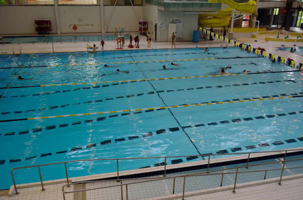 Großes Hallenbad mit schwimmenden Menschen, umgeben von Geländern und einem gefliesten Boden, Glasfenster im Hintergrund und Gegenstände auf der rechten Seite.