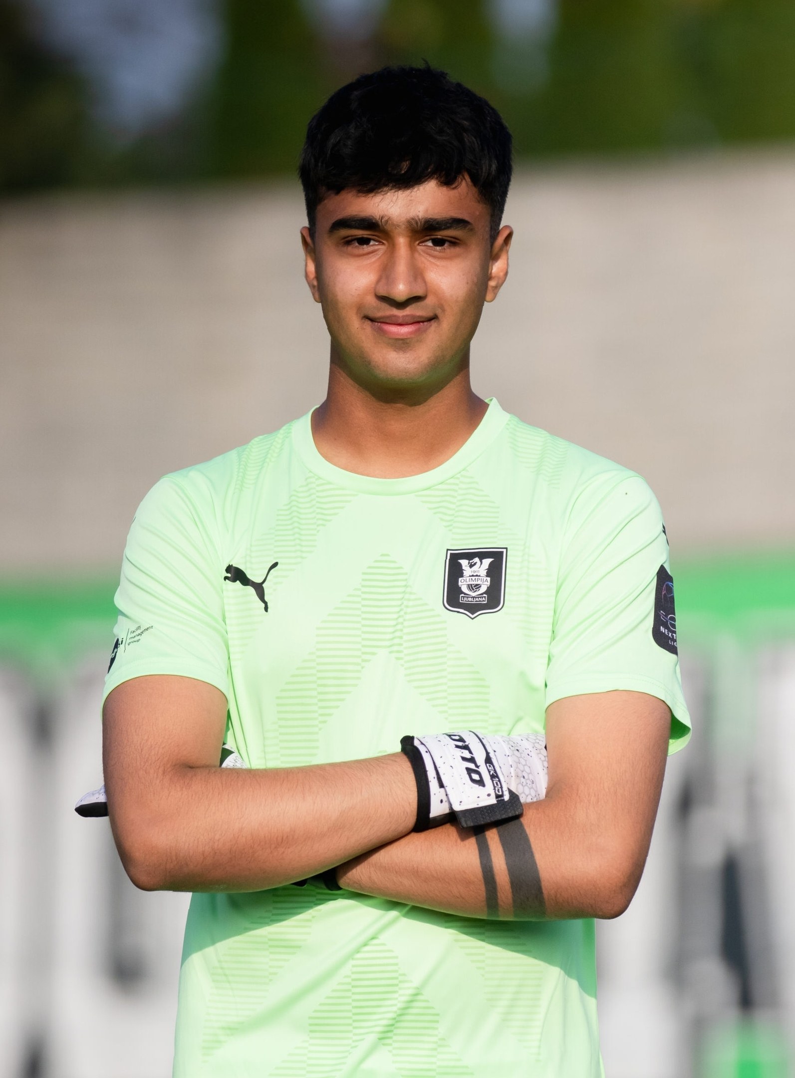 Ein junger Mann in einem grünen Hemd steht auf einem Fußballfeld und hat die Arme verschränkt, während er lächelt, wahrscheinlich ein Torwart, mit einem unscharfen Hintergrund.