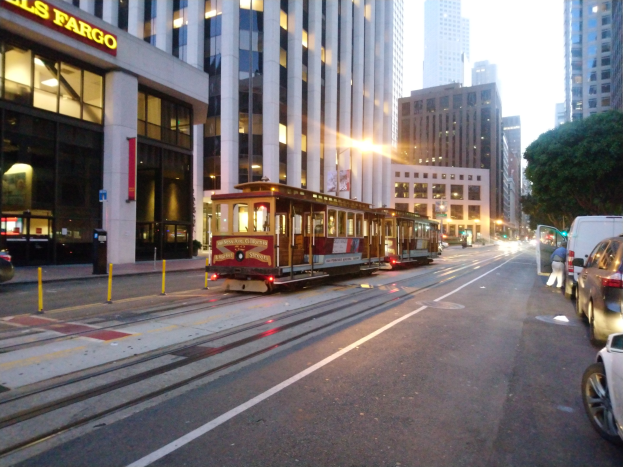 Eine Seilbahn fährt entlang einer städtischen Straße in San Francisco, mit Fahrzeugen, einem Fußgänger und einem geparkten Van, vor dem Hintergrund von Gebäuden, Bäumen und Himmel.