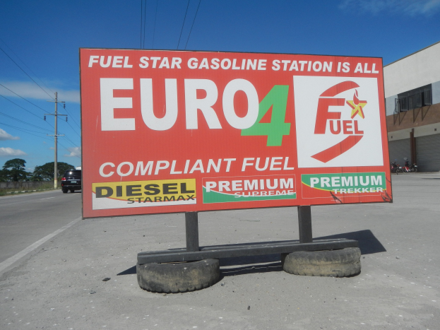 Eine Tankstelle mit einem "Euro4 Kraftstoff"-Schild im Vordergrund, Fahrzeuge auf der Straße, Strommästele mit Drähten, Bäume, ein Gebäude auf der rechten Seite und der Himmel überall.