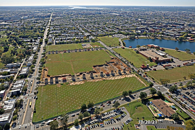 Luftaufnahme einer Stadt mit Gebäuden, Bäumen, Gras, Fahrzeugen, Wasser und Wolken, die einen großen zentralen Platz zeigt, der für einen Park vorgesehen ist.