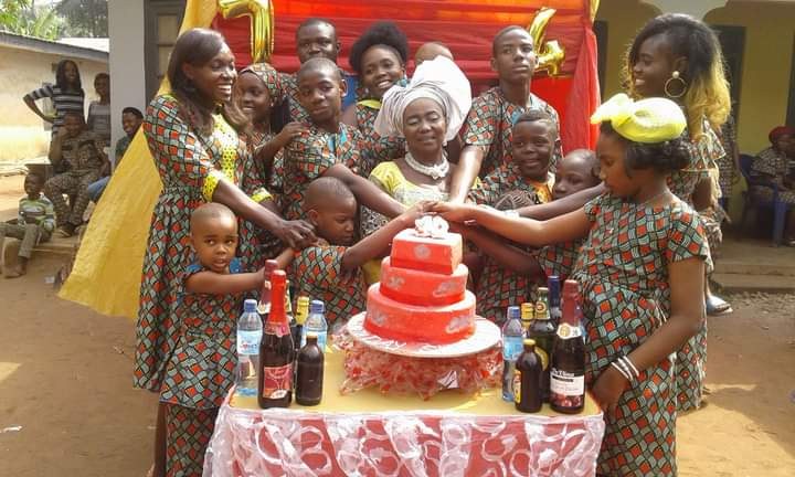 Eine Gruppe von Menschen sitzt um einen Tisch mit einem Kuchen, Flaschen und anderen Gegenständen in einer ländlichen Umgebung mit Häusern, Bäumen und einem klaren blauen Himmel, wahrscheinlich bei einer Geburtstagsfeier.