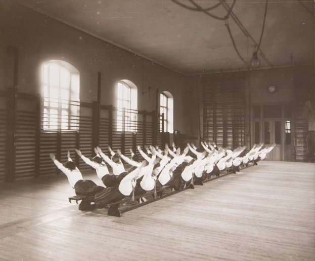 Gruppe von Menschen, die Pilates-Übungen in einem Fitnessstudio mit Fenstern und einer Uhr an der Wand durchführen.