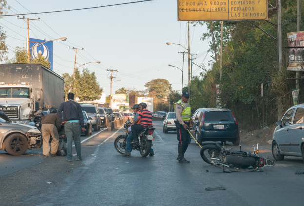 Eine Gruppe von Menschen steht um ein verunglücktes Motorrad auf der Straße mit mehreren Fahrzeugen, darunter ein Lastwagen, und einem Hintergrund aus Bäumen, Pfosten, Lampen und Schildern unter dem Himmel.