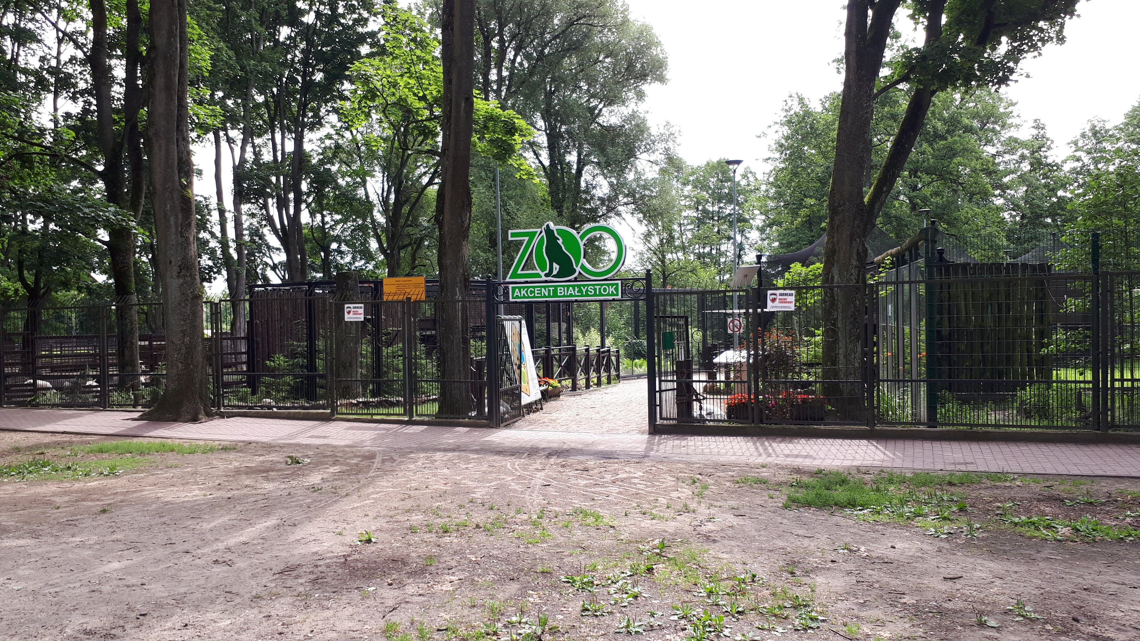 Eintritt zu einem Zoo mit einem "Zoo"-Schild, umgeben von einem Metallzaun, mit Bäumen, blühenden Pflanzen, einem Straßenpfahl und einem bewölkten Himmel.