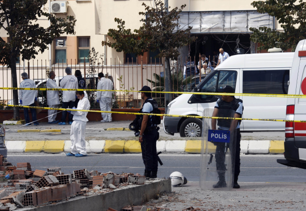 Eine Gruppe von Polizeibeamten steht vor einem Gebäude mit Fenstern, umgeben von Fahrzeugen, einem Zaun, Bäumen und Schutt, einschließlich Ziegelsteinen und einem Helm auf dem Boden, wahrscheinlich nach einer Bombenexplosion in Mexiko-Stadt.