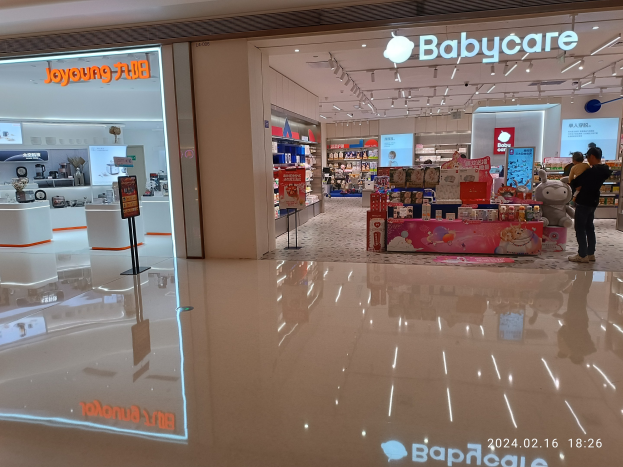 Ein Babyladen in einem Einkaufszentrum mit Menschen, Spielzeugen, Regalen, Schildern, einem Display, Deckenleuchten und einem Dach.
