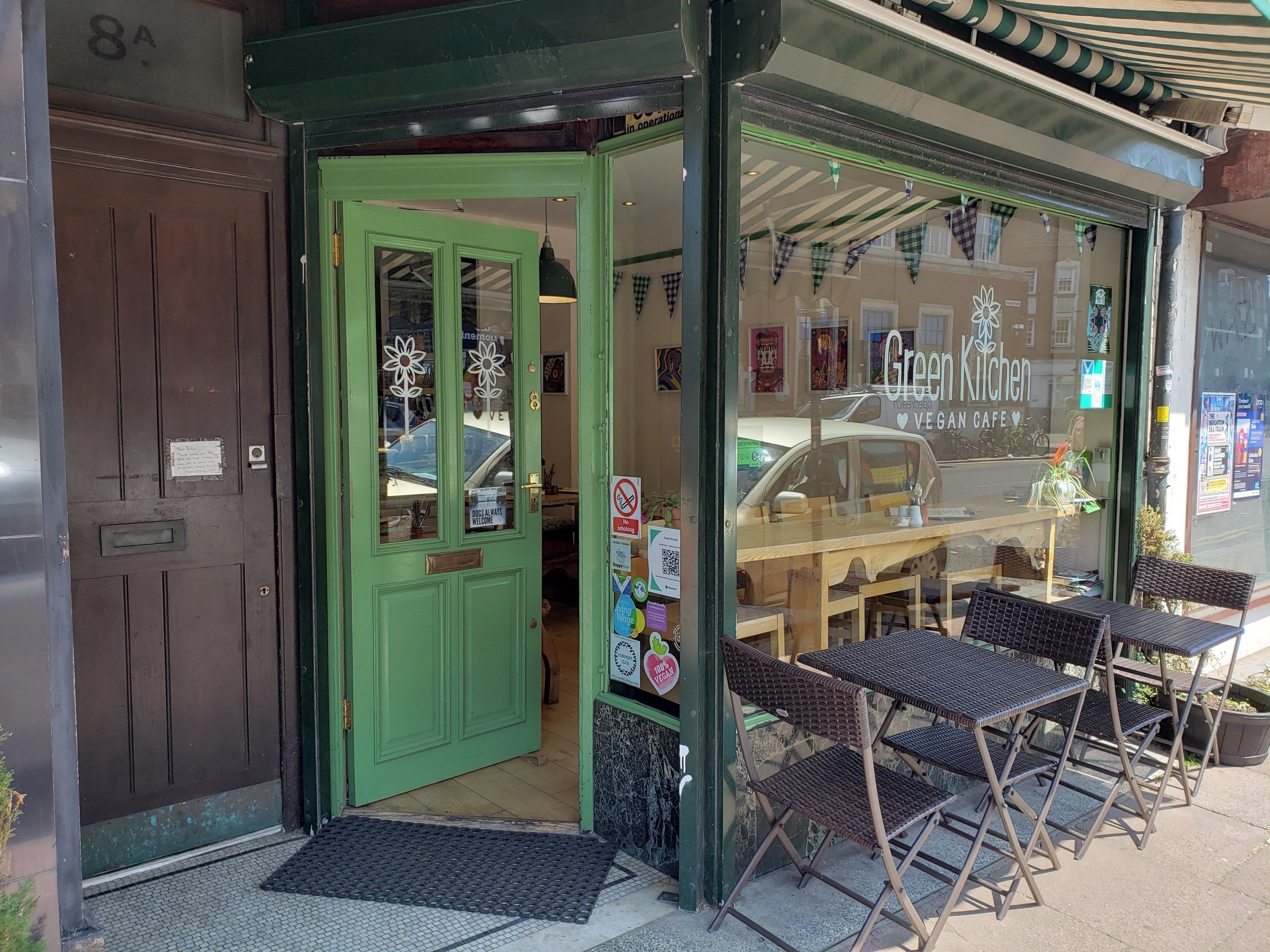 Außensitzbereich des Green Kitchen Vegan Cafe mit Tischen, Stühlen, Pflanzen, einer Fußmatte und einer Glastür mit Postern.