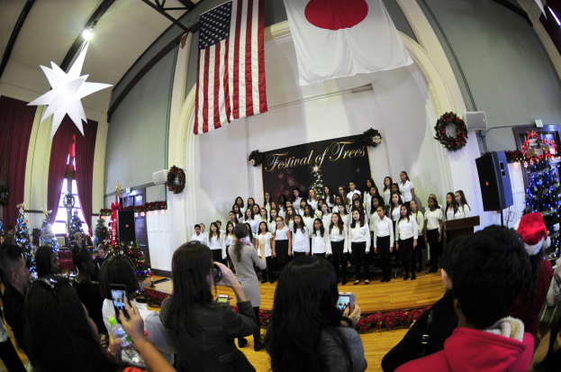 Eine Gruppe von Menschen auf einer Bühne mit Weihnachtsdekoration, einem Podium, Lautsprechern, einer Texttafel und einem Publikum mit Kameras und Handys.