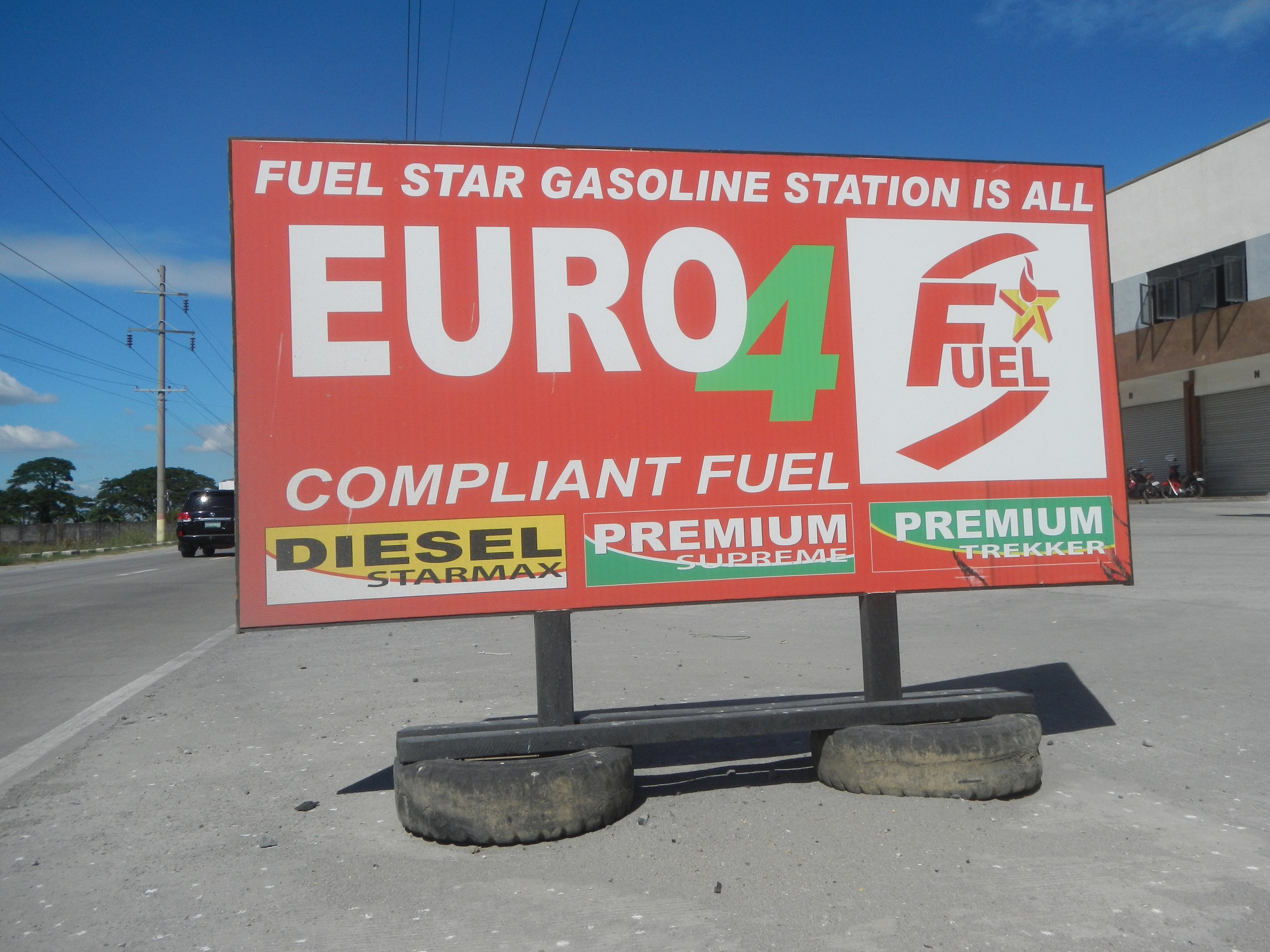 Eine Tankstelle mit einem "Euro4 Fuel"-Schild im Vordergrund, Fahrzeuge auf der Straße, Strommästele mit Drähten, Bäume und ein Gebäude im Hintergrund, der Himmel ist sichtbar.