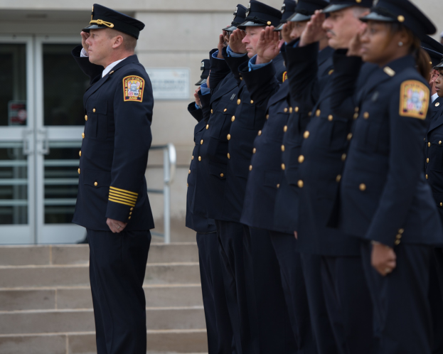 Gruppe von Polizisten in Uniform, die in Formation vor einem Gebäude mit Glastüren und Treppen salutieren.