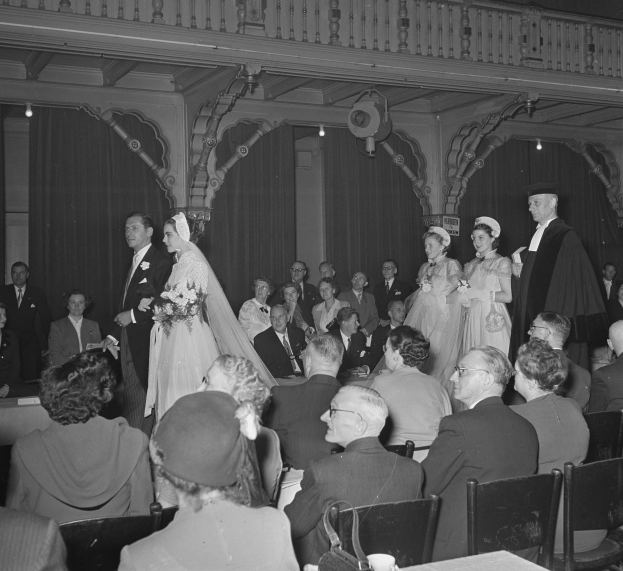 Schwarz-weißes Foto einer Hochzeitszeremonie in einem großen Raum mit sitzenden und stehenden Gästen, einige halten Blumensträuße, Tische mit Tassen und Gegenständen sowie Vorhänge und Deckenleuchten im Hintergrund.