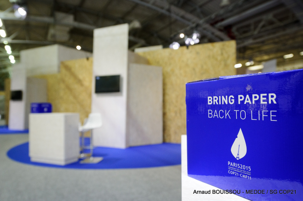 Messestände mit einem blauen und weißen Schild, auf dem "Bring Paper Back to Life" steht, auf der rechten Seite, mit unscharfen Stühlen, Tischen, einem Wandfernseher und Deckenleuchten im Hintergrund.