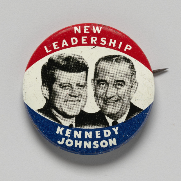 Weißer Ansteckbutton mit Abbildungen von Kennedy und Johnson und dem Text "Neue Führung" auf einer glatten Oberfläche.