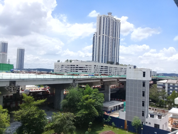 Luftaufnahme einer Stadtlandschaft mit Gebäuden, Bäumen, Gras, Fahrzeugen, einer Brücke, Masten und einem bewölktem Himmel, die eine Baustelle für eine neue Wohnanlage zeigt.