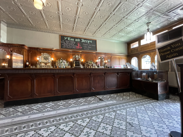 Ein großer Raum mit einer zentralen Bar, beleuchtet von Deckenlampen, mit Flaschen auf einem Tisch und einer Tafel mit Text auf der rechten Seite, im Tom und Jerry Pub in London.
