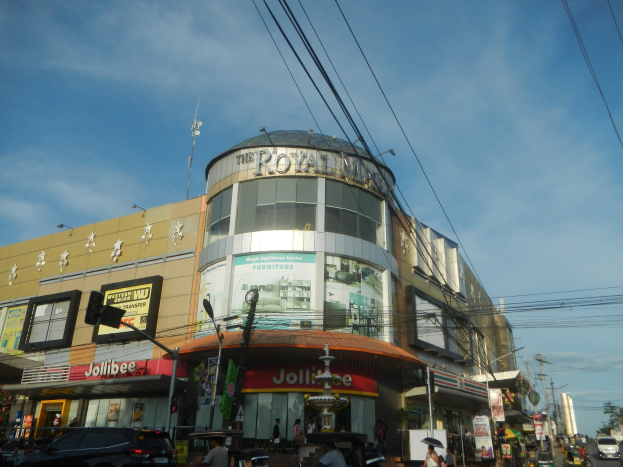 Eine belebte Stra├če mit Fahrzeugen, Passanten und dem Royal Mall Einkaufszentrum im Hintergrund, umgeben von B├Ąumen, Strommasten und Dr├Ąhten unter einem Himmel.