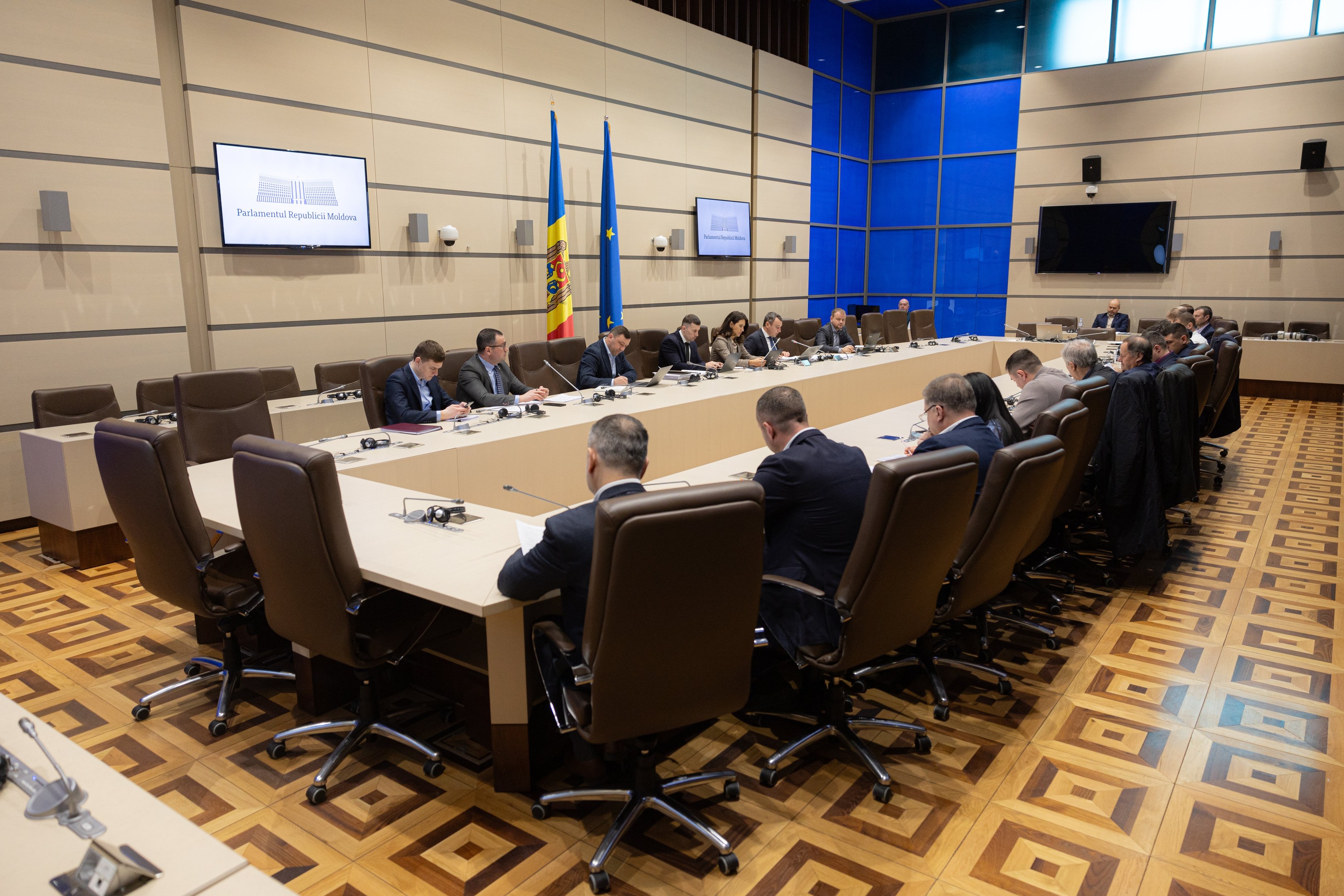Große Gruppe von Menschen um einen langen Tisch mit Mikrofonen und Papieren in einem Raum mit Flaggen der Europäischen Union, Fernsehern, Deckenleuchten und Lautsprechern.