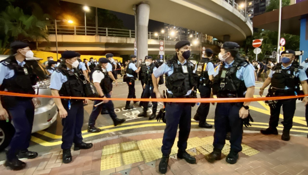 Polizeibeamte in Uniform und mit Masken stehen vor einer Menge, mit Fahrzeugen, Gebäuden, Bäumen und einer Brücke im Hintergrund, nach einer Demonstration in Hong Kong.