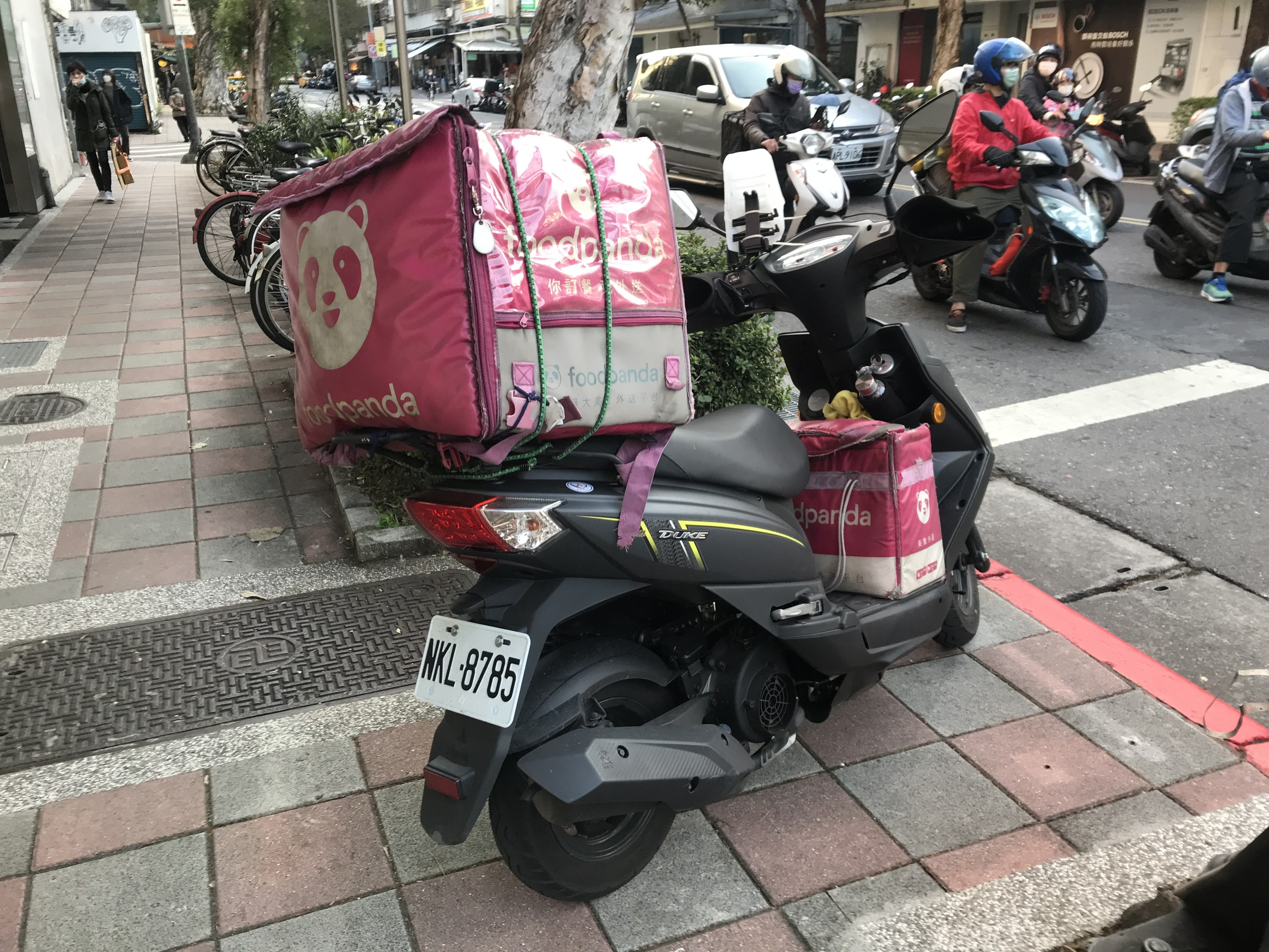 Ein Roller an der Straßenseite neben einem Gebäude geparkt, mit einem Auto, einem Fahrrad und Passanten in der Nähe und Bäumen, Gebäuden und Schildern im Hintergrund.