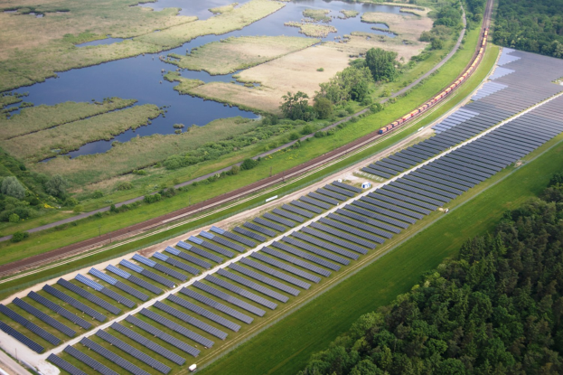 Luftaufnahme eines Solarparks mit Solarpanelen in einem Feld, umgeben von Bäumen, Gras, Pflanzen und Wasser, mit einem Zug auf einer Bahnstrecke.