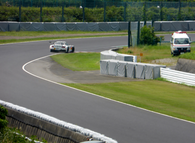 Ein Rennwagen fährt eine Rennstrecke entlang, im Hintergrund ist ein Polizeiauto zu sehen, umgeben von Grünflächen und Absperrungen, mit Text am unteren Bildrand.