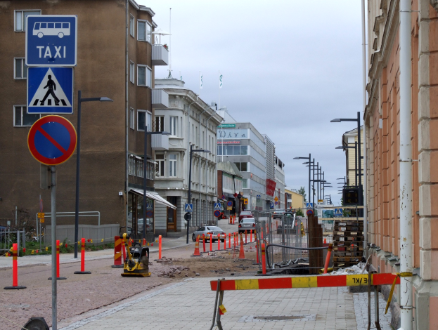 Stadtstraßenszene mit Gebäuden, Straßeninfrastruktur, Fahrzeugen, einer Baustelle und Bäumen gegen einen bewölkten Himmel.