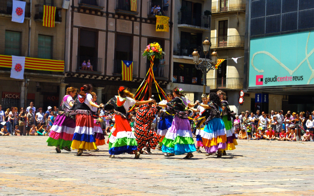 Eine Gruppe von Frauen in bunten Kleidern tanzt auf der Straße, umgeben von einer Menge Menschen, mit Gebäuden, Gelöndern, Balkonen, Flaggen, einem Laternenmast und einer Tafel mit Schrift im Hintergrund.