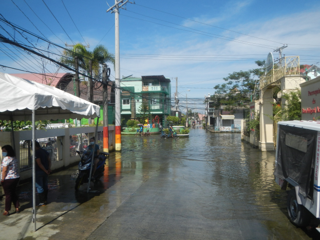 Überschwemmte Stadtstraße mit Fahrzeugen, Fußgängern, einem Zelt, Versorgungsmasten, Gebäuden, Bäumen und bewölktem Himmel.