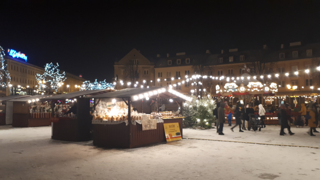 Ein nächtlicher Weihnachtsmarkt in einer verschneiten Straße mit Menschen, Kinderwagen, beleuchteten Hütten, Pflanzen, Bäumen, Gebäuden, Schildern und einem bewölkten Himmel.