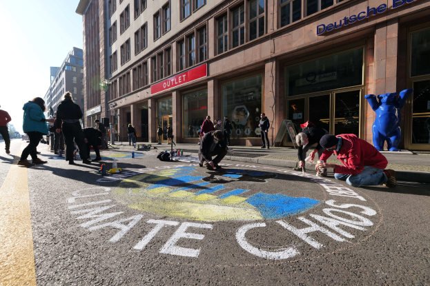 Menschen sitzen vor einem Gebäude mit Fenstern und Namensschildern, umgeben von Flaschen und anderen Gegenständen, während sie an einer Klimawandel-Demonstration in Berlin teilnehmen, die von Bäumen und einem klaren blauen Himmel umgeben ist.