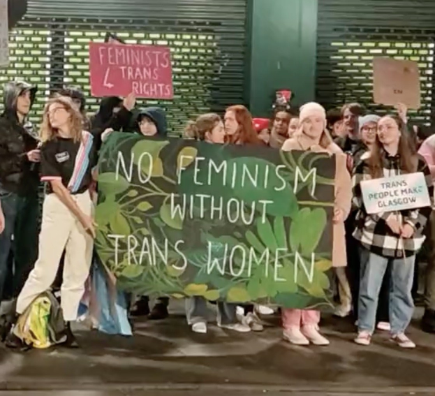 Eine Gruppe von Menschen mit einem Banner "Kein Feminismus ohne Transfrauen" und verschiedenen Schildern in einem öffentlichen Raum.