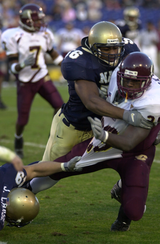 Zwei Fußballspieler in Helmen und Uniformen kämpfen auf dem Feld, während Teamkollegen und Zuschauer im Hintergrund zusehen.