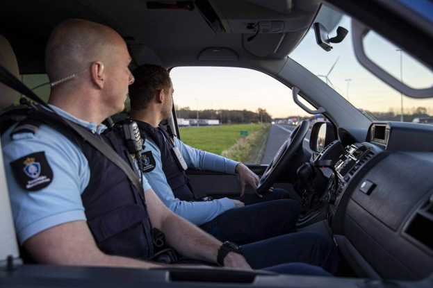 Zwei Polizisten sitzen in einem Auto auf einer Straße mit Gras, Bäumen und Polen unter einem klaren blauen Himmel.