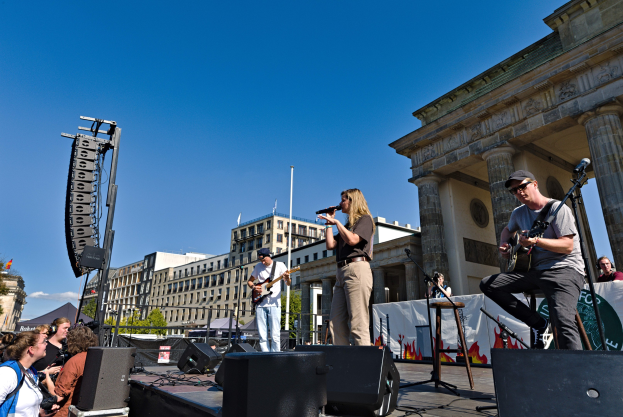 Eine Gruppe von Menschen, die auf einer Bühne vor dem Brandenburger Tor in Berlin Musik machen, umgeben von Lautsprechern und Equipment, vor einer Kulisse aus Gebäuden, Bäumen und einem klaren Himmel.