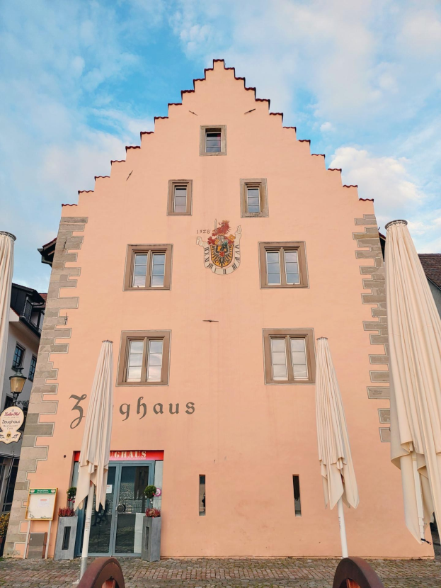 Ein pinkes Gebäude mit einem zentralen Uhrenturm, umgeben von Fenstern, Schirmen, einem Schild, einer Straßenlaterne, Topfpflanzen und einem bewölkten Himmel in München, Deutschland.