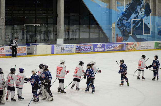 Gruppe junger Menschen, die Eis Hockey auf einer Indoor-Eisfläche spielen, mit Helmen und Hockey-Schlägern ausgestattet, mit einer Person auf der linken Seite und Bannern, Säulen und Glaswänden im Hintergrund.