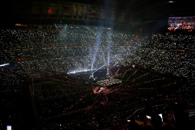 Eine große Menschenmenge in einem hell erleuchteten Stadion bei Nacht, mit einem Bildschirm auf der rechten Seite, der die Ergebnisse der Super Bowl XLVII-Halbzeitshow anzeigt.