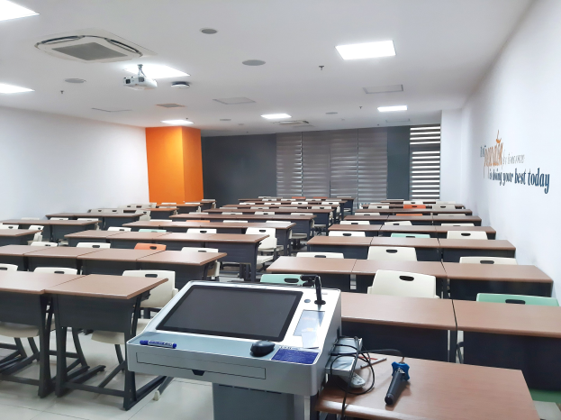 Ein Klassenzimmer mit Tischen und Stühlen in Reihen, einen Deckenprojektor und Gegenstände auf Tischen, mit Text an der rechten Wand, der auf eine Privatschule in Singapur hinweist.