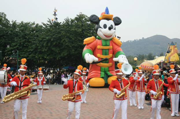 Eine Gruppe von Menschen in Kostümen spielt Musikinstrumente in der Nähe eines Mickey-Maus-Floats während eines Umzugs, mit Bäumen, Gebäuden, Hügeln und einem klaren blauen Himmel im Hintergrund und einer Tafel mit Text auf der linken Seite.