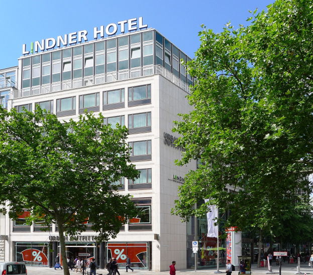 Großes modernes Hotelgebäude mit Glasfenstern, sichtbarer Beschriftung, umgeben von Bäumen und einer belebten Straße mit Fußgängern und Fahrzeugen unter einem klaren Himmel.